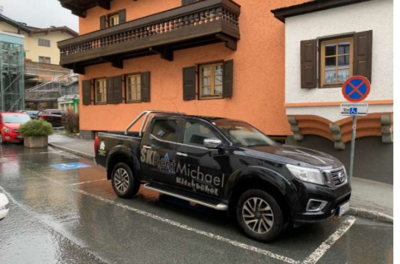 Behindertenparkplatz - Graggaugasse 5 Ein schwarzer Pickup mit der Aufschrift "SKI Michael" parkt an einer Straße in einem malerischen Dorf. Im Hintergrund sind traditionelle Gebäude und ein muffiger Himmel zu sehen.