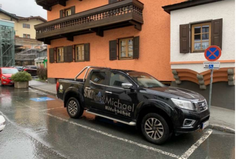 Behindertenparkplatz - Graggaugasse 5 Ein schwarzer Pickup mit der Aufschrift "SKI Michael" parkt an einer Straße in einem malerischen Dorf. Im Hintergrund sind traditionelle Gebäude und ein muffiger Himmel zu sehen.