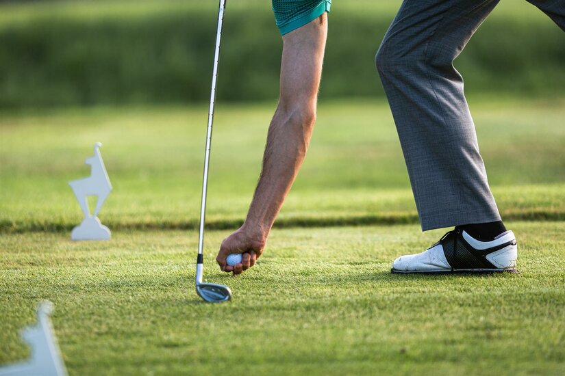 Golfschule Ein Golfer bereitet sich darauf vor, den Ball zu schlagen. Im Hintergrund sind Golfmarkierungen auf dem Platz sichtbar.