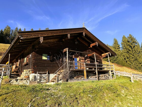 Eine charmante Almhütte aus Holz, umgeben von üppigem Grün und Bergen. Der Himmel ist blau mit einigen Wolken.