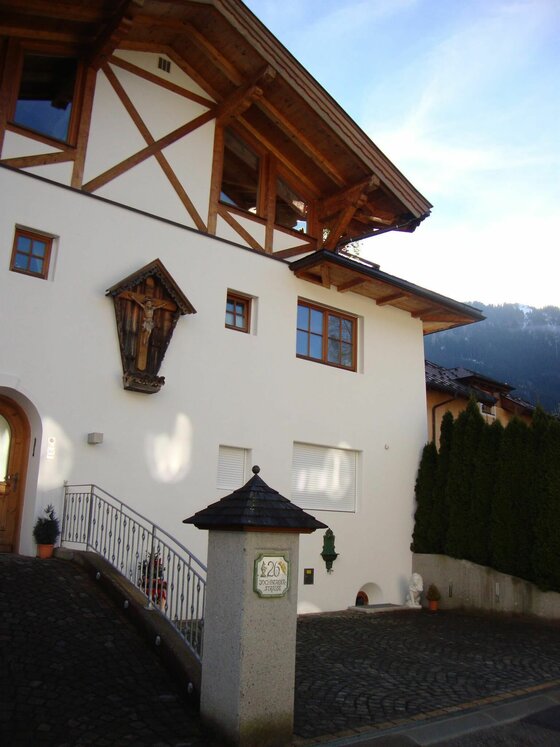 Haus Andreas in Kitzbühel Vorderansicht eines weißen Hauses mit Holzgiebel, Kruzifix an der Fassade und gepflasterter Einfahrt.