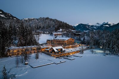 Beleuchtetes Hotel am verschneiten Seeufer vor bewaldeten Bergen in der Dämmerung.
