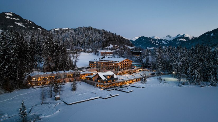 Beleuchtetes Hotel am verschneiten Seeufer vor bewaldeten Bergen in der Dämmerung.