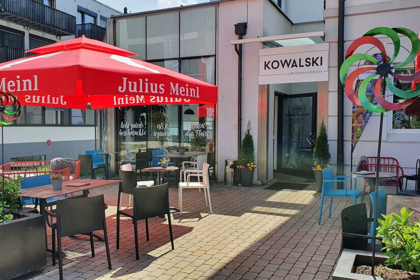 Ein einladendes Café mit roten Sonnenschirmen und bunten Stühlen. Die Terrasse ist hell und freundlich mit Pflanzen und einem Spielrad.