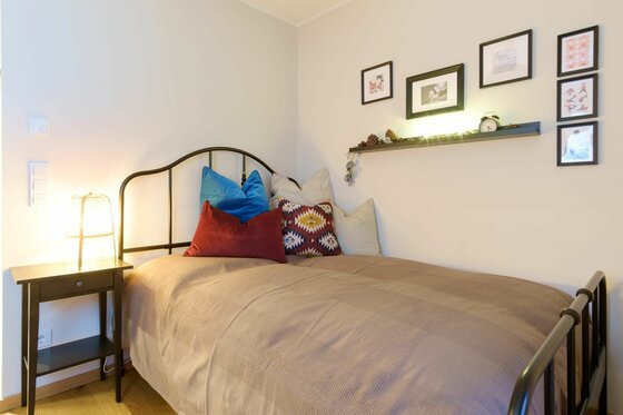 Einzelbett mit Metallrahmen, bunten Kissen, Nachttisch und Wanddekoration in einem hellen Zimmer.