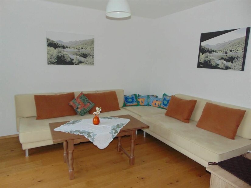 Künstler Ap. 1 Wohnraum Wohnzimmer mit Ecksofa, Holztisch und Landschaftsbildern an weißen Wänden.