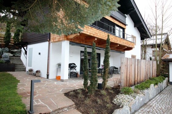 Modernes alpines Wohnhaus mit weißer Fassade, Holzschindeln am Balkon und einer gepflasterten Terrasse.