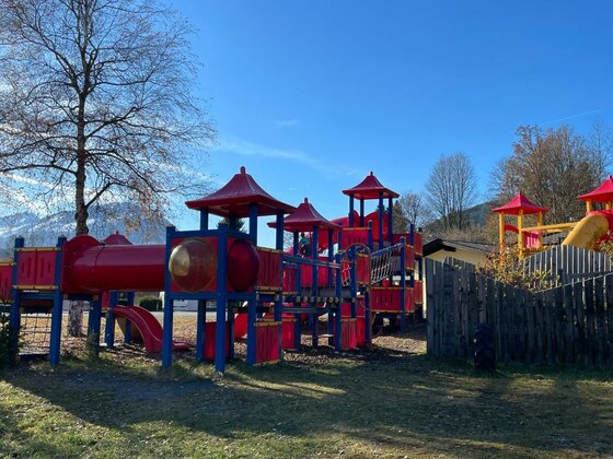 Spielplatz am Campingplatz Großes rotes Spielgerüst mit Türmen, Rutschen und Röhren auf einer Wiese vor Bergen.