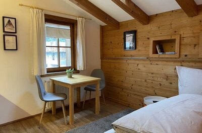 Innenansicht der kleinen Wohnung Zimmer mit Holzwänden, Bett, Holztisch, zwei grauen Stühlen und einem Fenster.