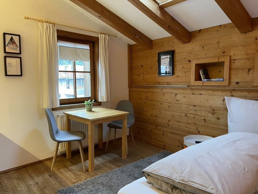 Innenansicht der kleinen Wohnung Zimmer mit Holzwänden, Bett, Holztisch, zwei grauen Stühlen und einem Fenster.