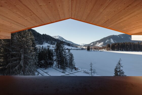 Blick von einem Holzunterstand auf einen verschneiten See und bewaldete Berge bei klarem Himmel.