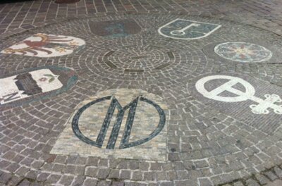 Stadtwappen der Schwesternstädte Eine dekorative Pflasterung mit verschiedenen Wappen und Symbolen. Die Gestaltung zeigt kunstvolle Muster auf dem Boden.