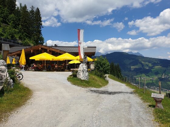 Berghütte mit gelben Sonnenschirmen auf einer Terrasse neben einem Schotterweg vor Gebirgspanorama.