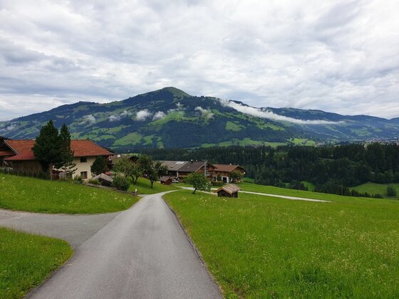 Asphaltierter Weg führt durch grüne Wiesen zu einer kleinen Siedlung vor bewaldeten Bergen unter bewölktem Himmel.