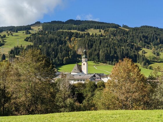 Blick auf die Pfarrkirche Jochberg vor bewaldetem Berghang unter blauem Himmel.
