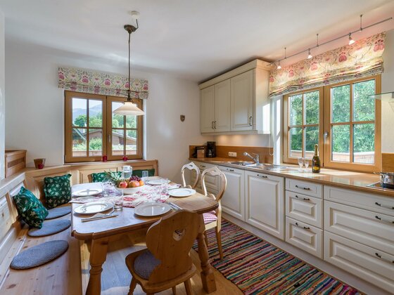 Helle Landhausküche mit Holztisch, Eckbank und weißer Küchenzeile vor zwei Fenstern.