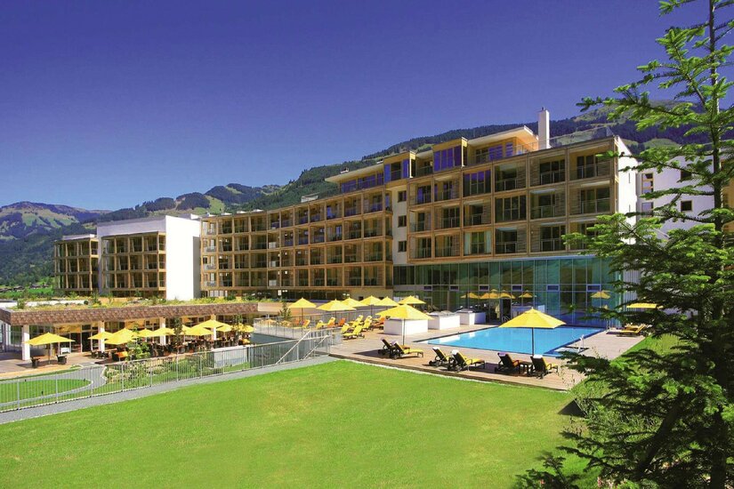 Modernes Hotelgebäude mit Außenpool, gelben Sonnenschirmen und Liegewiese vor bewaldeten Bergen.