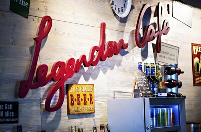 Legenden Cafe in Kitzbühel Roter Schriftzug Legenden Cafe an einer Holzwand mit Getränkeflaschen und Schildern.