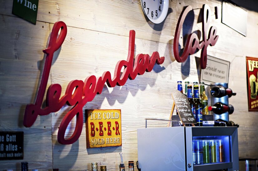 Roter Schriftzug Legenden Cafe an einer Holzwand mit Getränkeflaschen und Schildern.