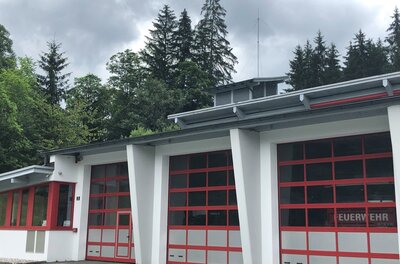 Feuerwehr <p>Ein modernes Feuerwehrgebäude mit roten Garagentoren in Aurach. Im Hintergrund sind hohe Bäume und graue Wolken zu sehen.</p>