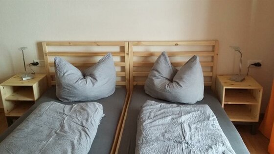 Zwei nebeneinanderstehende Holzbetten mit grauer Bettwäsche und Nachttischen.