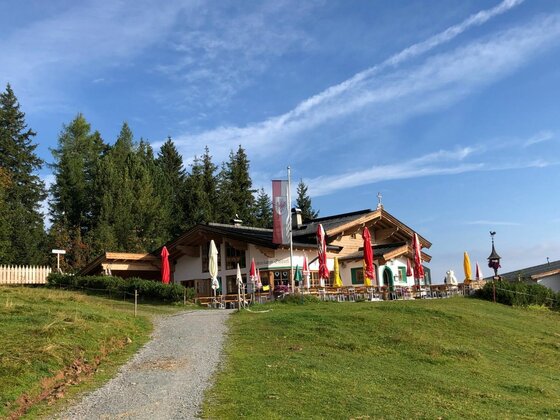 Berghütte an der Hüttenrunde Berghütte mit Außenterrasse und Sonnenschirmen vor Nadelwald unter blauem Himmel.
