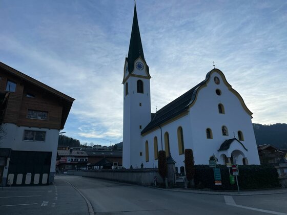 Kirche am Radweg Wilder Kaiser Weiße Dorfkirche mit spitzem Turm und geschwungenem Giebel an einer asphaltierten Straße.