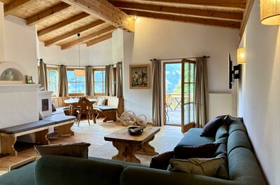 IMG_1904 2 Ein einladendes Wohnzimmer mit Holzbalkendecke und großen Fenstern. Gemütliche Sitzgelegenheiten und ein Esstisch schaffen eine warme Atmosphäre.
