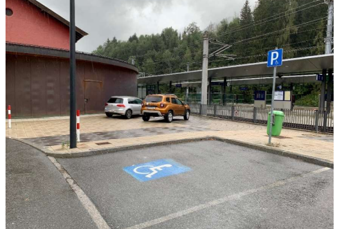 Behindertenparkplatz - ÖBB Hauptbahnhof Kitzbühel Ein Parkplatz mit zwei Autos, darunter ein behindertengerechter Parkplatz. Im Hintergrund ist eine Bahnhofsplattform und Bäume zu sehen.