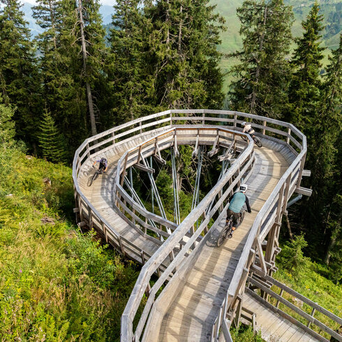 Eine geschwungene Holzbrücke am Sonnenrast Trail in Kitzbühel, umgeben von üppigem Grün, während Mountainbiker die Strecke nutzen.