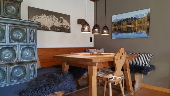 Rustikale Sitzecke mit Holztisch, Eckbank, Kachelofen und drei Hängelampen in einer Ferienwohnung.