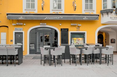 Außenansicht der Bar Jimmy's mit gelber Fassade, grauer Markise und Stehtischen im Außenbereich.