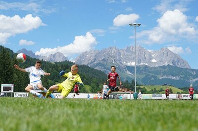 Kinder spielen Fußball auf einem Rasenplatz vor einer alpinen Bergkulisse.