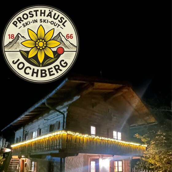 Ein traditionelles Holzhaus mit schöner Beleuchtung in der Nacht. Oben ist ein Logo mit einer Blume und den Worten "Prosthäusl Jochberg" zu sehen.