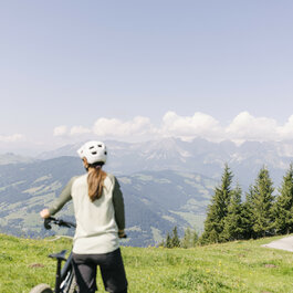 Eine Radfahrerin steht auf einer Wiese am Hahnenkamm in Kitzbühel und blickt auf die umliegenden Berge und das grüne Tal.