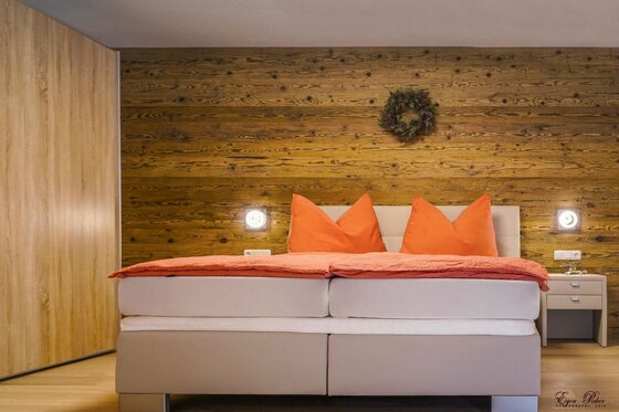 Doppelbett mit orangefarbener Bettwäsche vor einer rustikalen Holzwand mit Kranz.