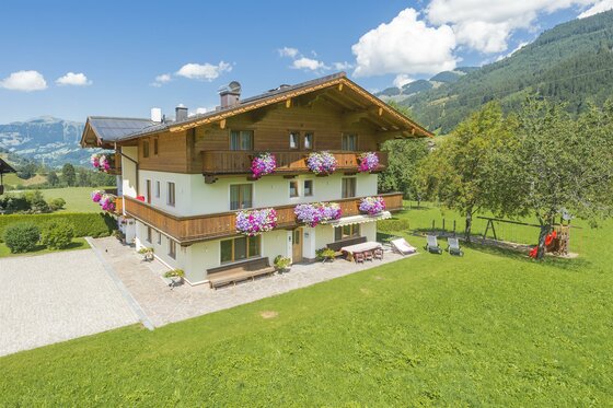 Dreistöckiges alpines Gästehaus mit Holzbalkonen, Blumenschmuck und Gartenanlage vor Bergpanorama.