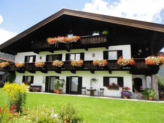 Alpines Landhaus mit weißer Fassade, dunklen Holzbalkonen und üppigem Blumenschmuck vor einer Rasenfläche.