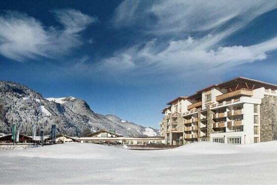 Grand Tirolia Kitzbühel Außenansicht Mehrstöckiges Hotelgebäude mit Steinfassade und Holzbalkonen in einer verschneiten Alpenlandschaft.