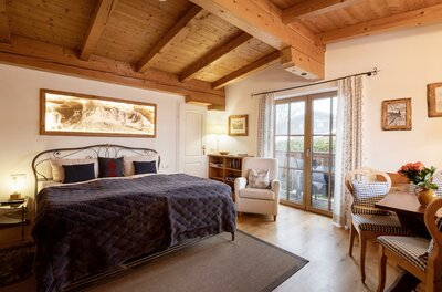 Ein gemütliches Schlafzimmer mit einem großen Bett und Holzbalkendecke. Es gibt einen kleinen Essbereich und einen Balkon mit Blick nach draußen.