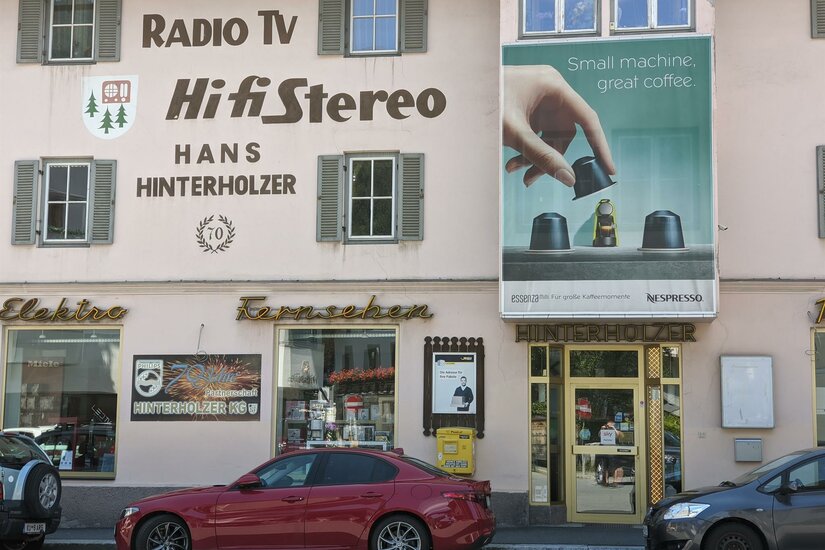 Hinterholzer Ein Geschäft mit einem großen Schild für HiFi-Geräte und Elektrotechnik. Nebenan ist eine Werbung für Nespresso-Kaffeemaschinen.