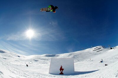 Ein Snowboarder springt über ein großes Hindernis im Schnee, während die Sonne hell am Himmel scheint. Die Landschaft ist schneebedeckt und die Umgebung ist klar und sonnig.