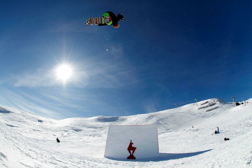 Ein Snowboarder springt über ein großes Hindernis im Schnee, während die Sonne hell am Himmel scheint. Die Landschaft ist schneebedeckt und die Umgebung ist klar und sonnig.