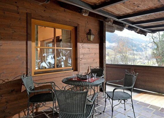 Terrasse Appartement Holzleitner Jochberg Überdachte Holzterrasse mit runden Metalltisch und vier Flechtstühlen vor einer Holzwand mit Fenster.