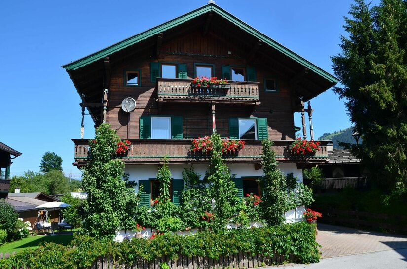 Haus Pircher Dreistöckiges Holzhaus mit Blumenbalkonen und grünen Fensterläden vor blauem Himmel.