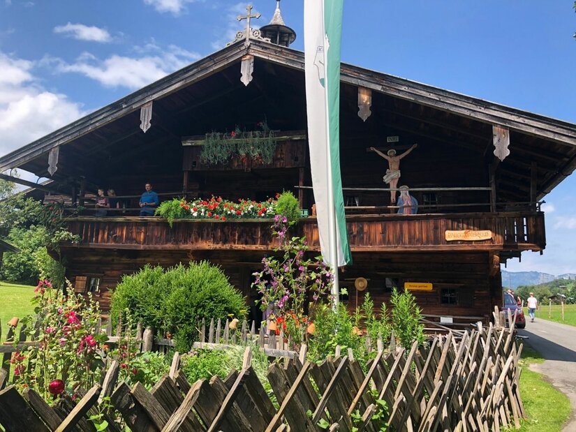 Traditionelles Bauernhaus aus dunklem Holz mit Blumenschmuck am Balkon und einem Holzzaun im Vordergrund.