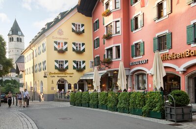Blick auf farbige Häuserfassaden mit Restaurants und einem Kirchturm im Hintergrund in einer gepflasterten Straße.