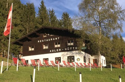 Zweistöckiges Holzgebäude mit weißem Erdgeschoss, roten Liegestühlen davor und einer österreichischen Flagge.