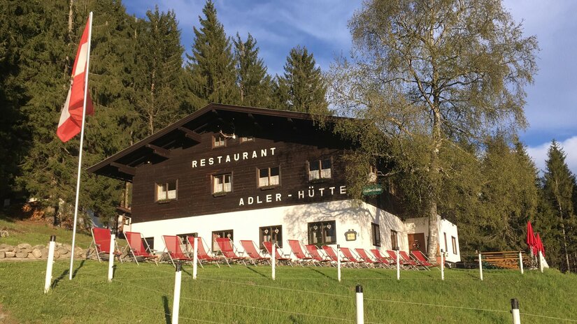 Adlerhütte im Sommer Zweistöckiges Holzgebäude mit weißem Erdgeschoss, roten Liegestühlen davor und einer österreichischen Flagge.
