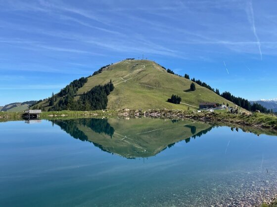 Ein grasbewachsener Berggipfel spiegelt sich in der glatten Oberfläche eines Speicherteichs unter blauem Himmel.